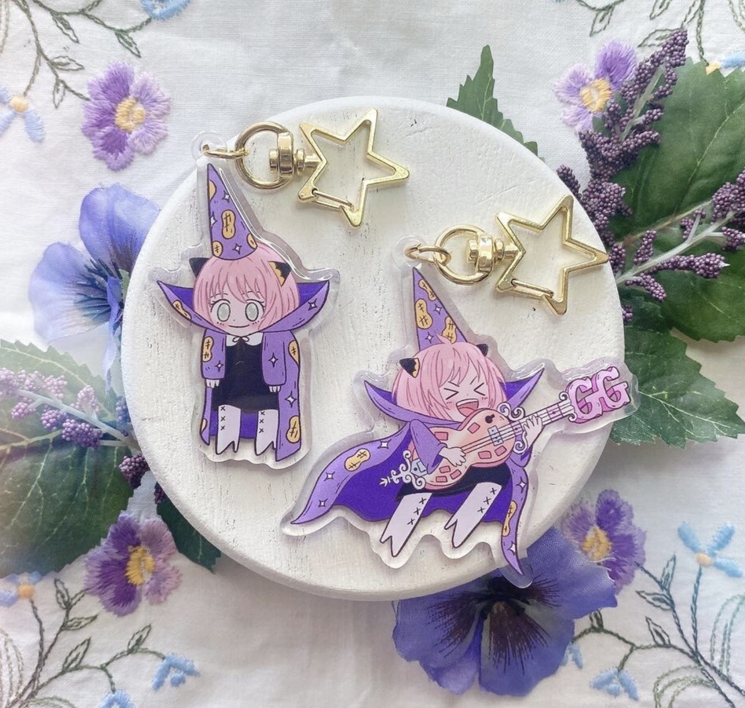 Two anime-style keychains with a star and witch designs, placed on a floral background