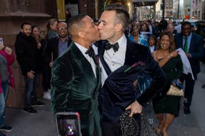 Two men in formal attire sharing a kiss, spectators smiling in the background