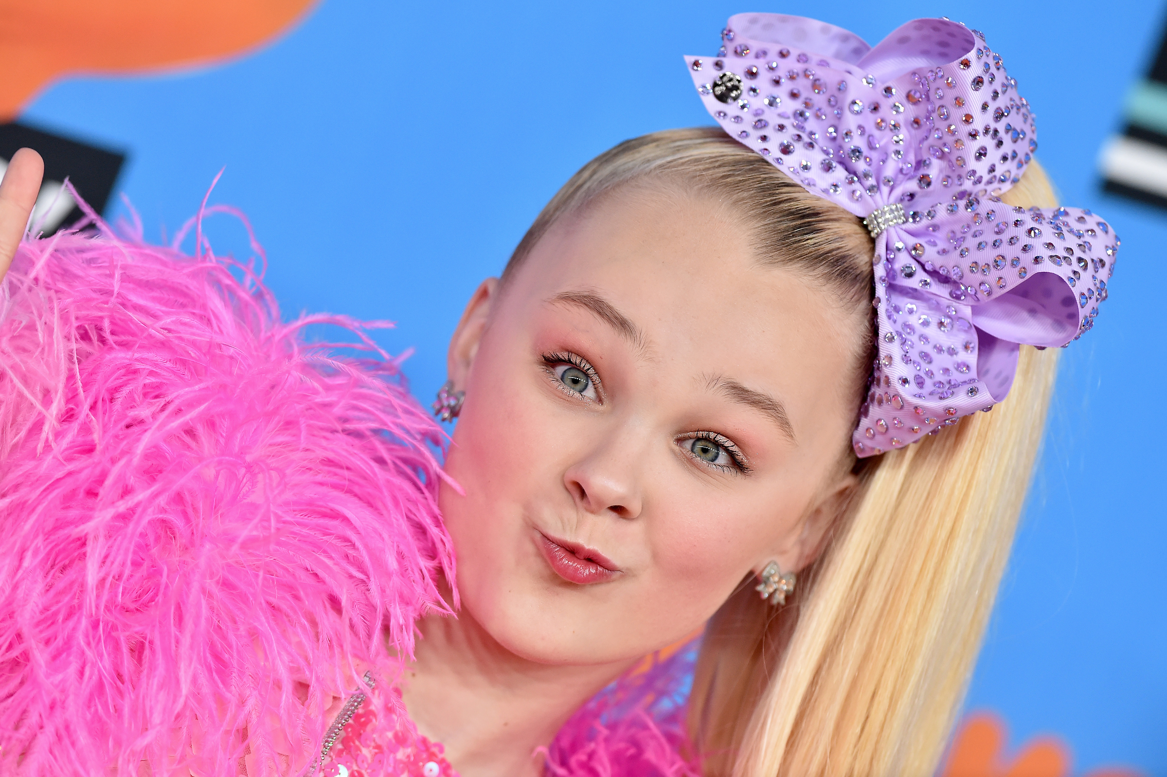 JoJo Siwa poses with a large purple bow, pink fringed outfit, pouting at the camera