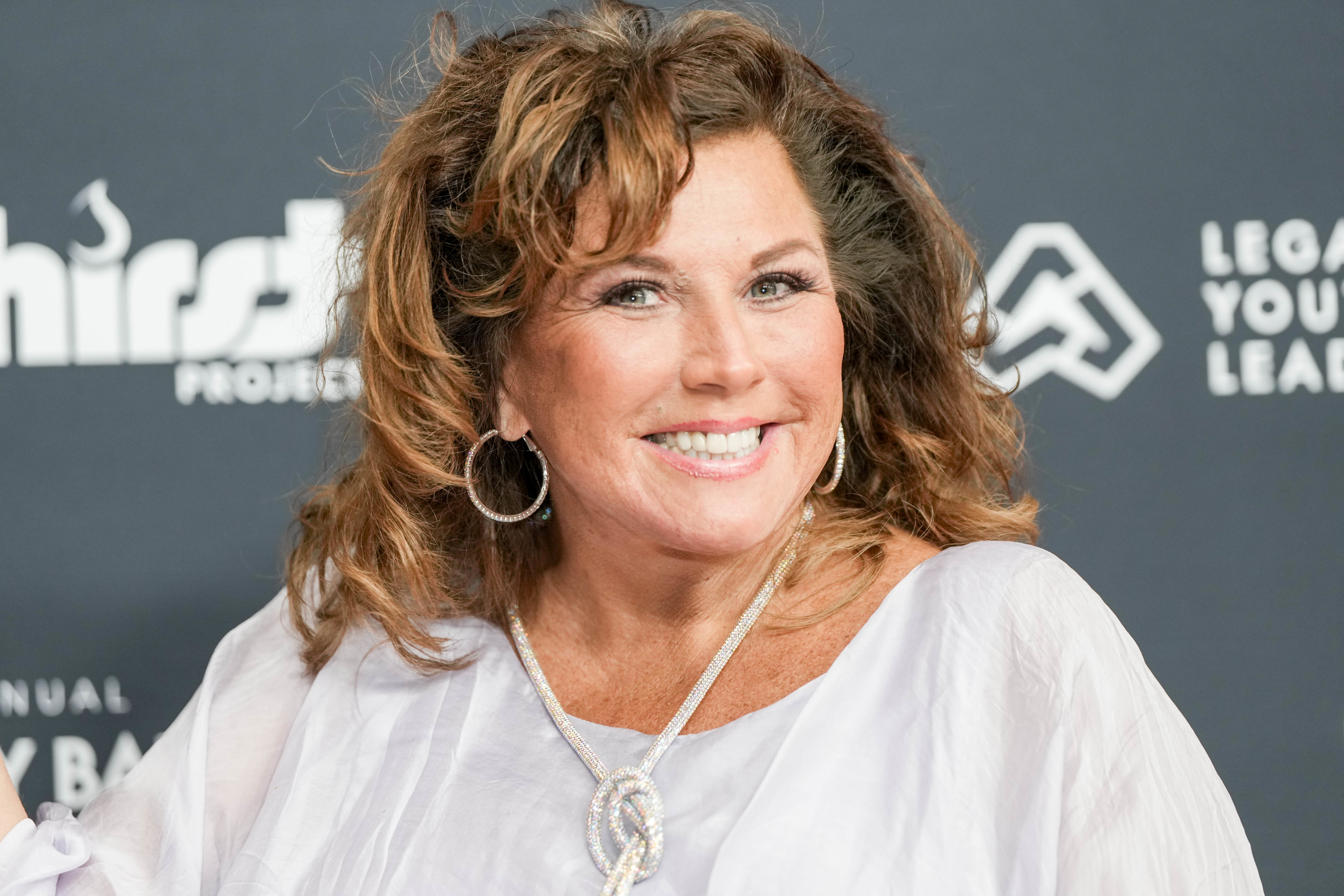 Abby Lee Miller smiling in a white blouse with large earrings and a necklace