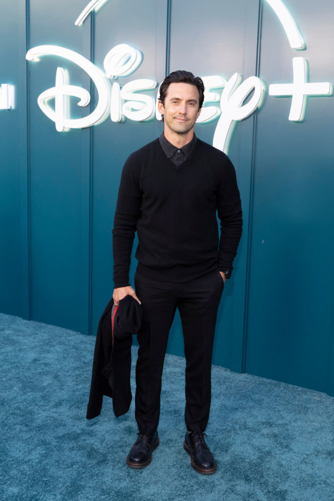 Milo Ventimiglia posing in a black sweater and trousers, holding a jacket, at a Disney+ event