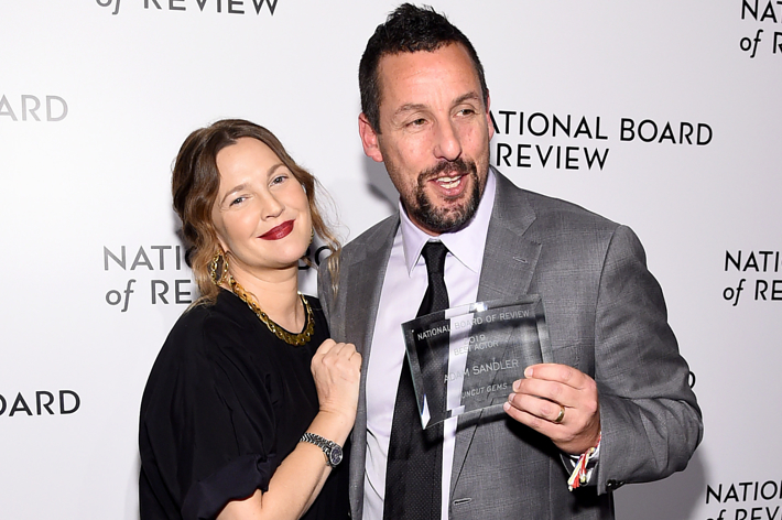 Adam Sandler and guest smiling, holding an award at a formal event. He's in a suit, she's in a dress and necklace.
