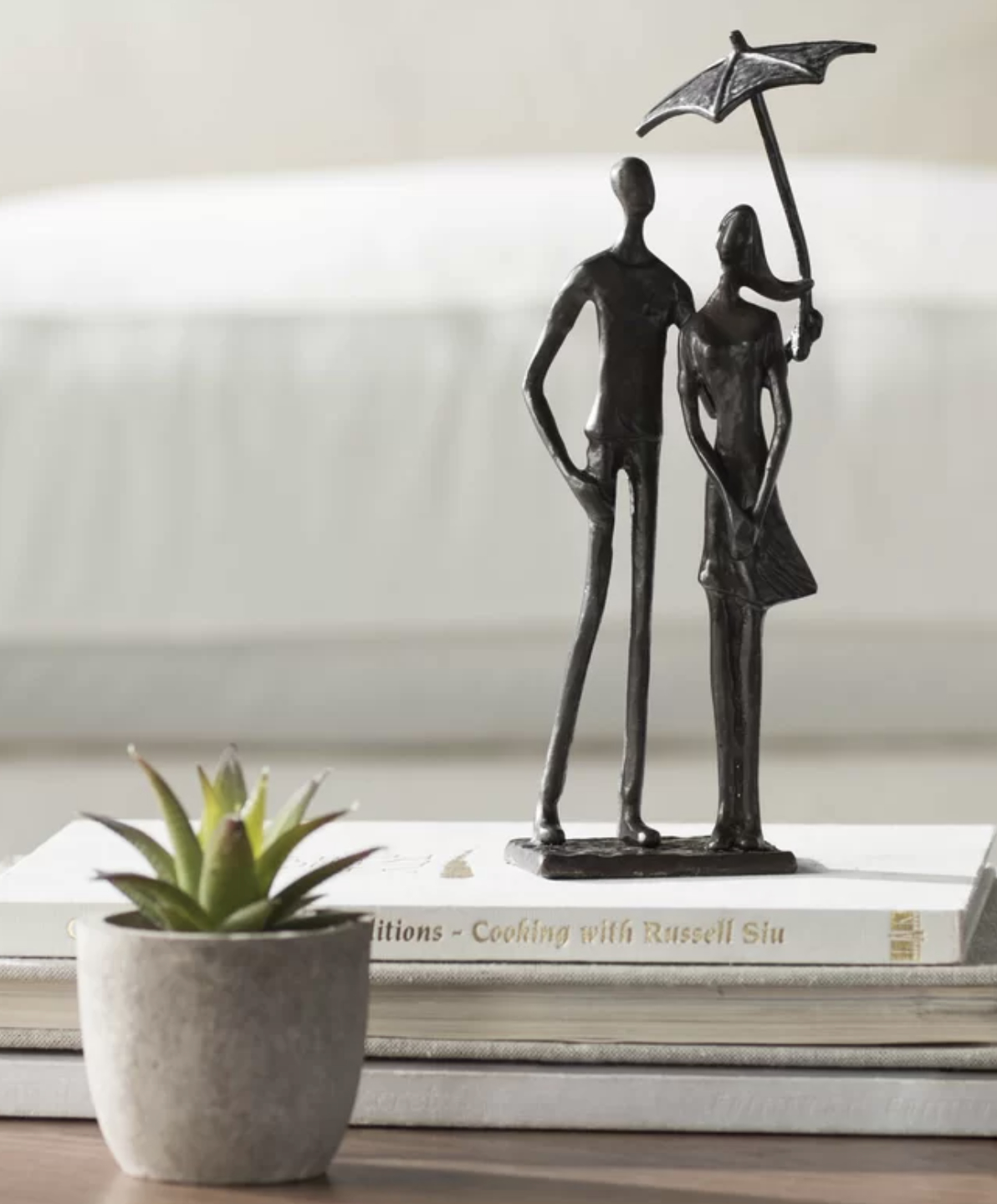 Sculpture of two abstract human figures, one holding an umbrella, on a book next to a potted plant