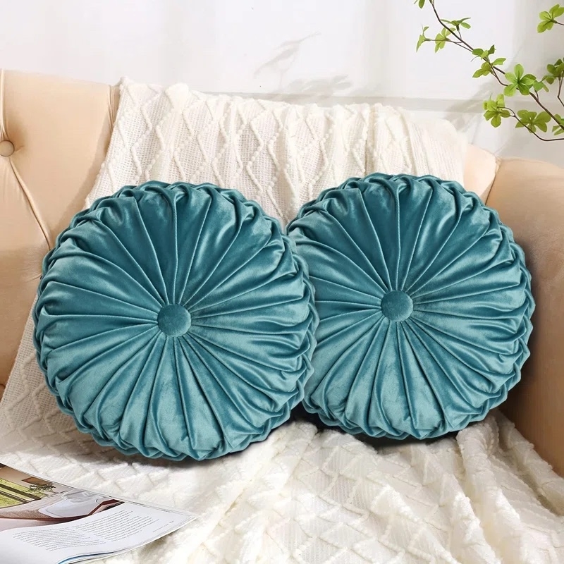 Two round, pleated velvet throw pillows on a couch with a knit blanket and magazine