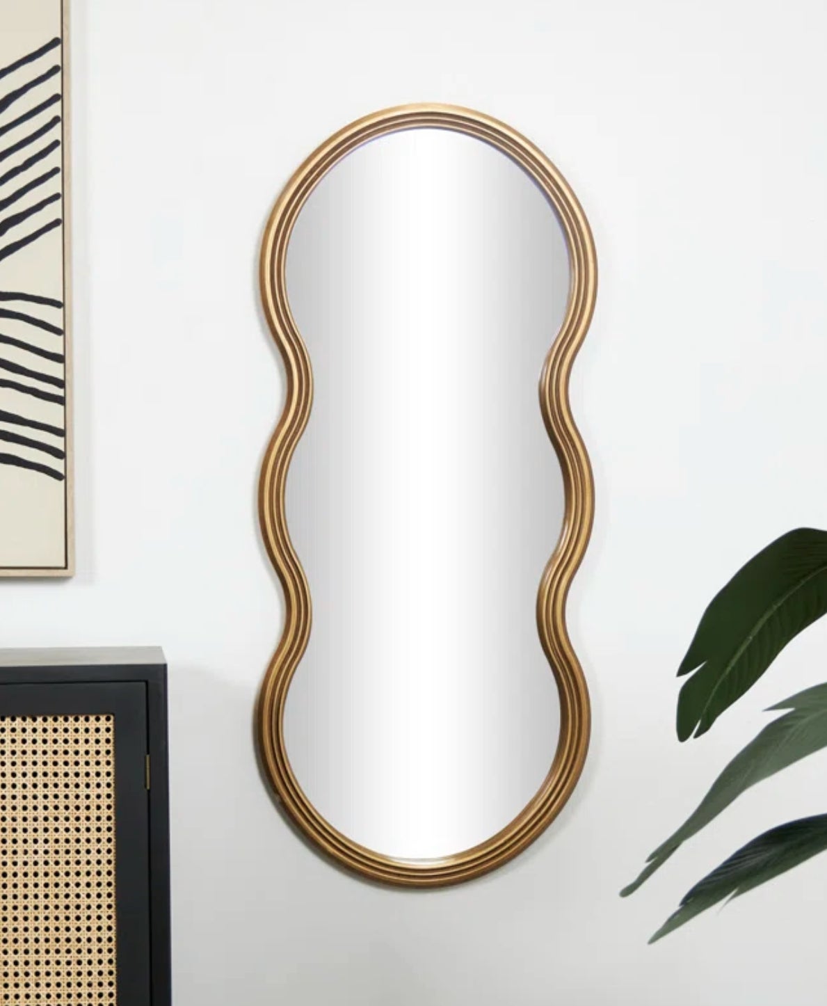 Oval wall mirror with a gold-tone frame, mounted beside artwork and above a black cabinet