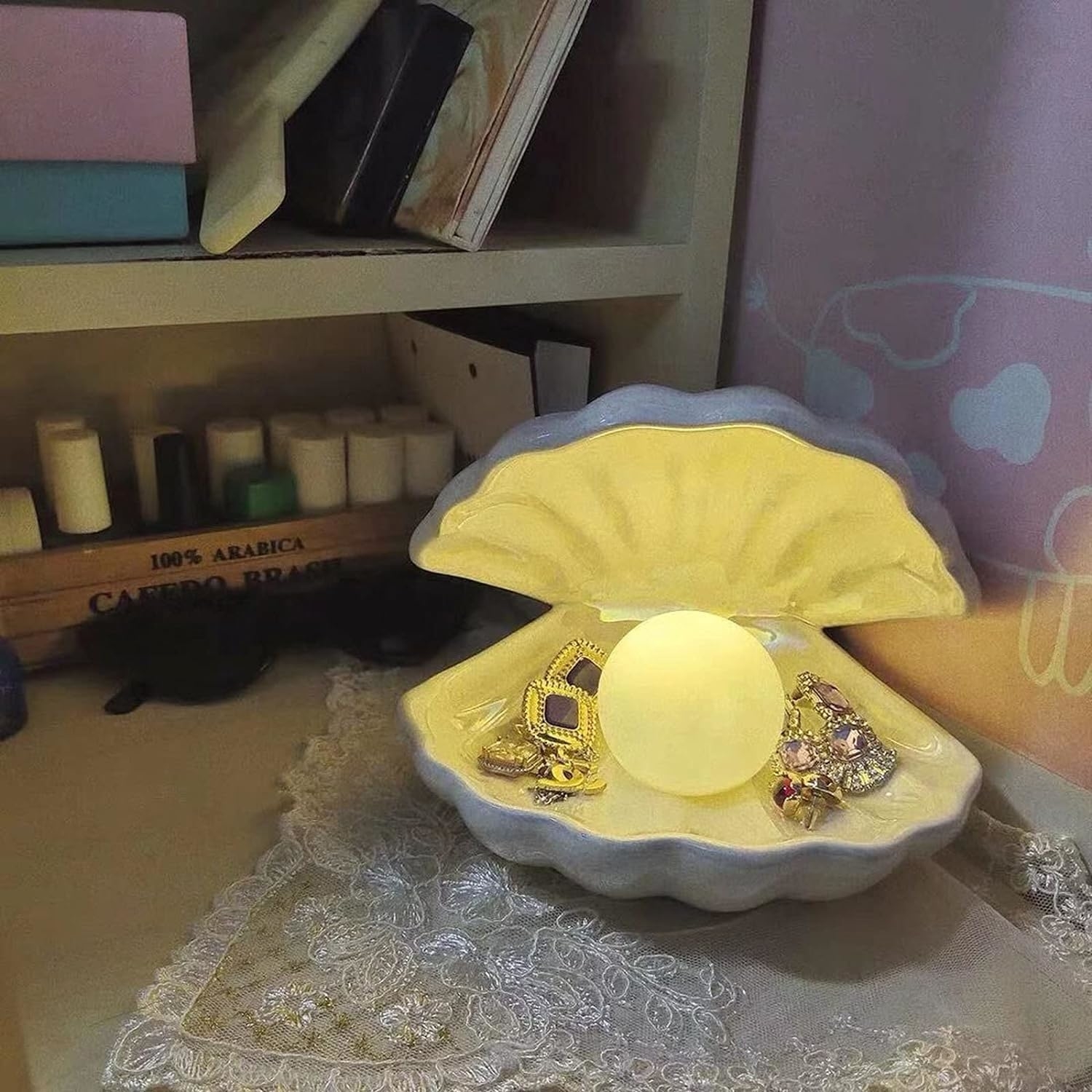 A shell-shaped jewelry dish with assorted jewelry and a spherical lamp, placed on a table with books in the background