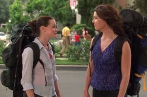 Two women facing each other with backpacks, one in a vest and blouse, the other in a sleeveless top
