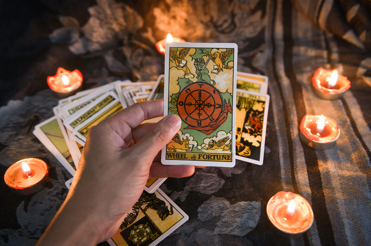 Hand holding a 'Wheel of Fortune' tarot card above a spread of other cards, surrounded by lit candles