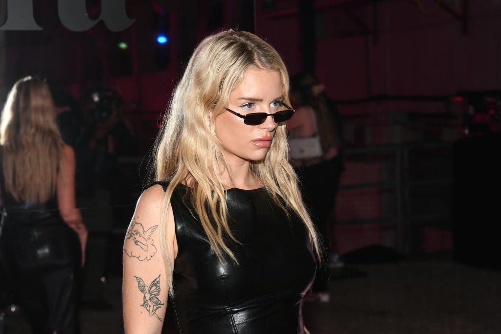 Woman in sleeveless black dress and sunglasses at an event, with visible tattoos on her arm