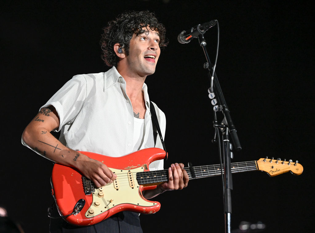 Man performing on stage with a guitar; in a white shirt, tattoos visible on arms