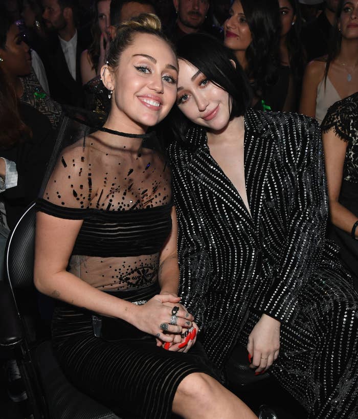 Miley Cyrus and Noah Cyrus sitting side by side, Miley in a sheer black top, Noah in a black beaded outfit