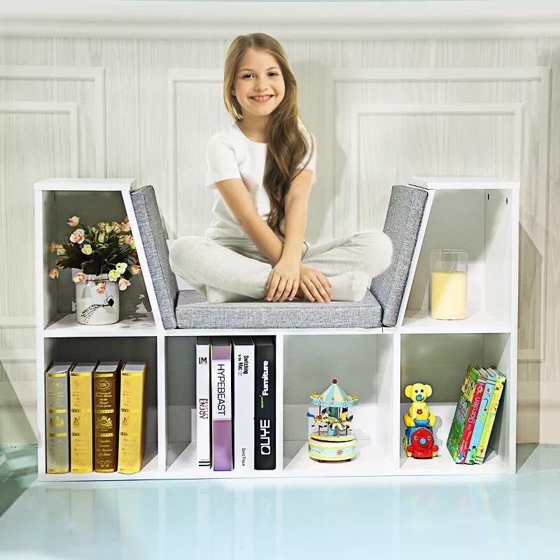 Smiling child sitting on a cushioned white bookshelf with books, toys, and a glass of juice