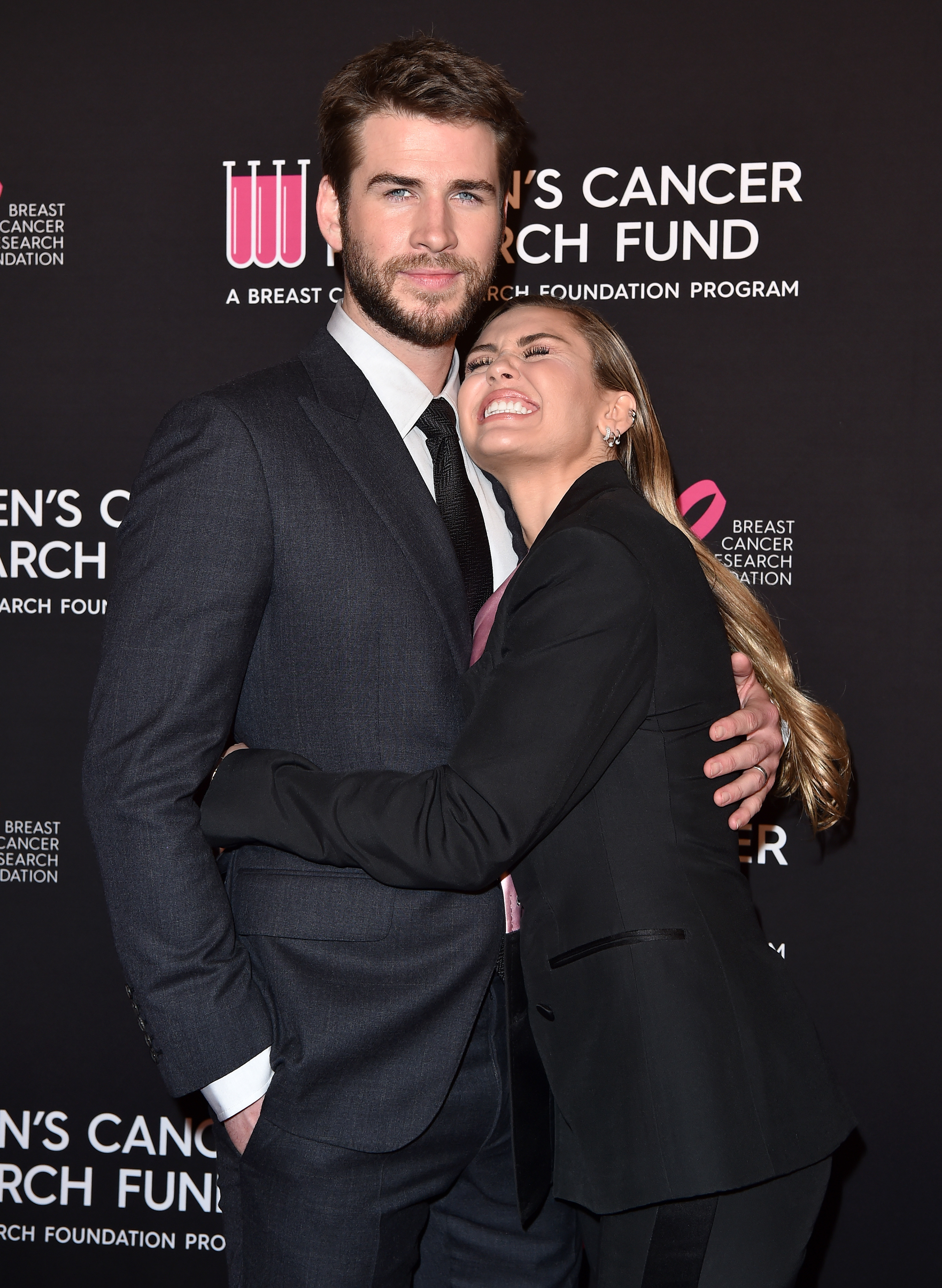 Liam Hemsworth in a suit hugging Miley Cyrus wearing a blazer at an event