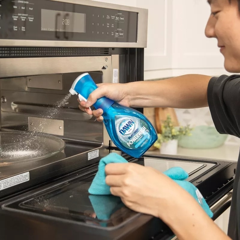 Person cleaning stove with spray bottle and cloth. Product shown for effective cleaning in the kitchen