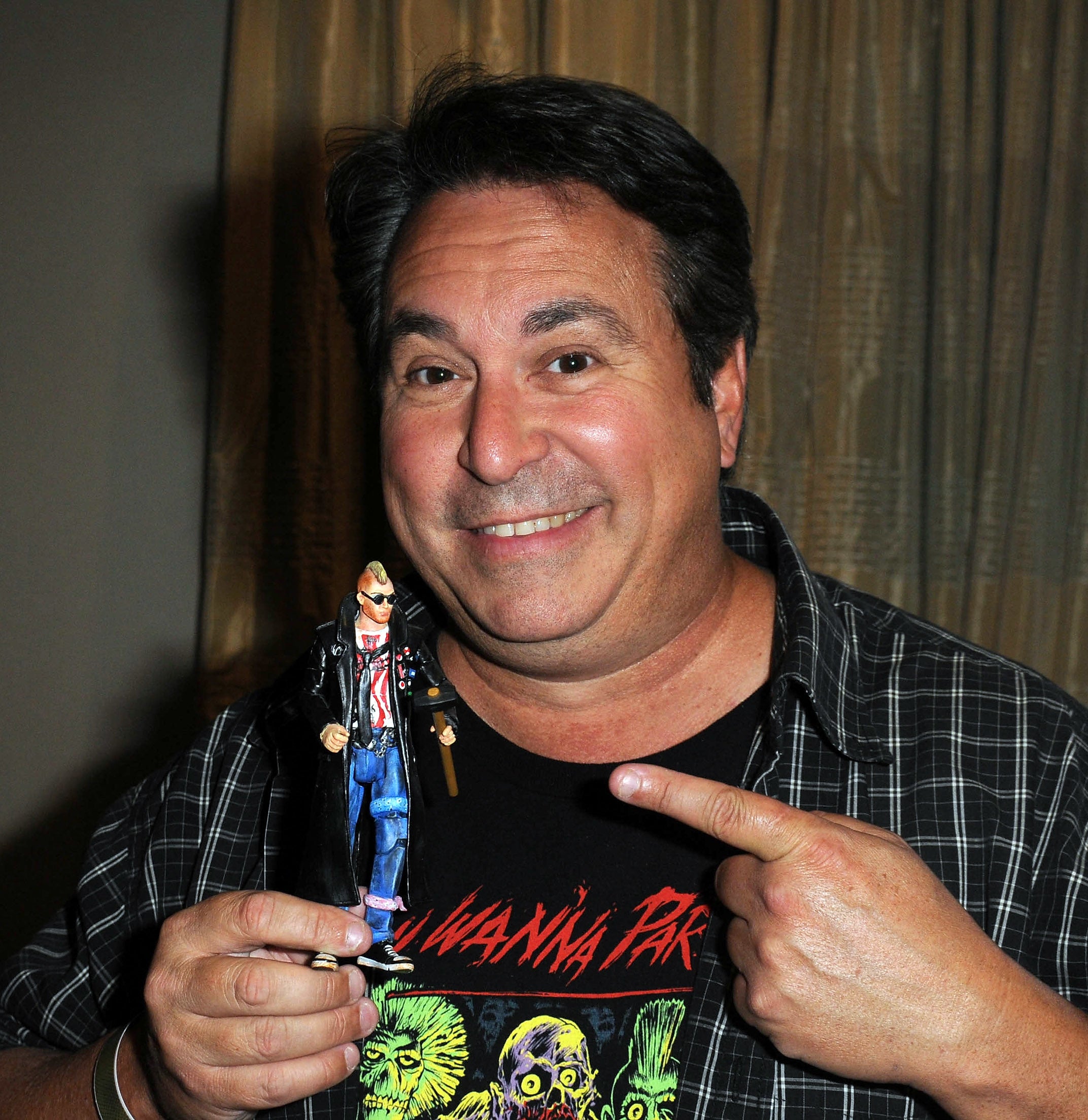 Man holding an action figure, pointing at it with a smile; he's wearing a graphic tee and checkered shirt