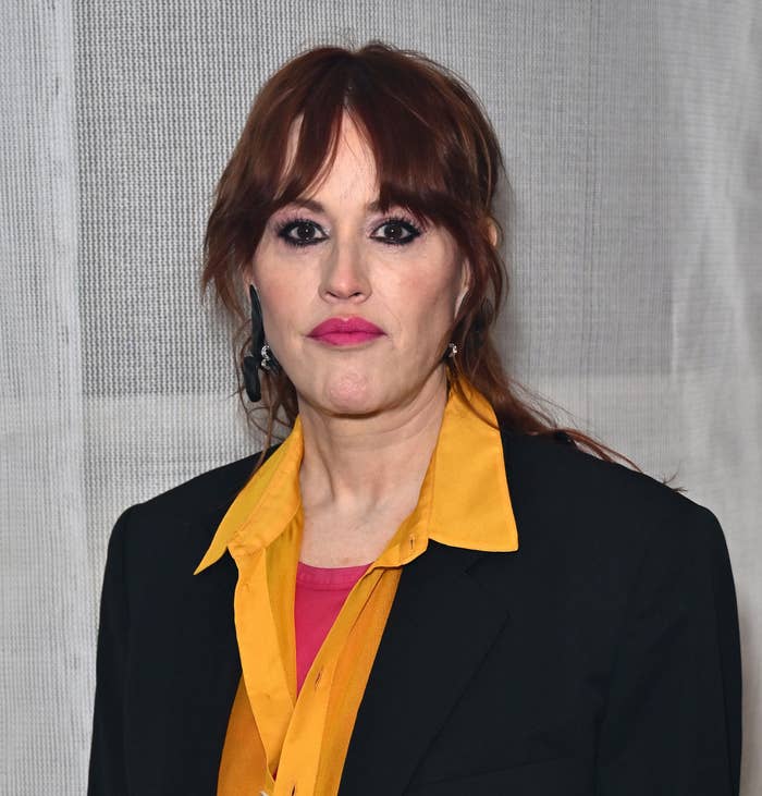 Molly Ringwald in a yellow blouse and black blazer standing against a neutral backdrop