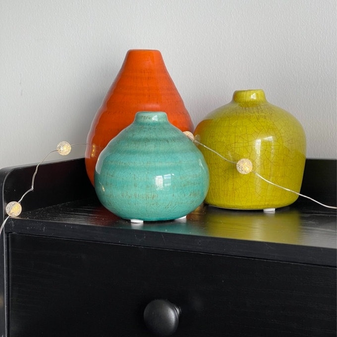 three ceramic vases on a dresser with a string of lights
