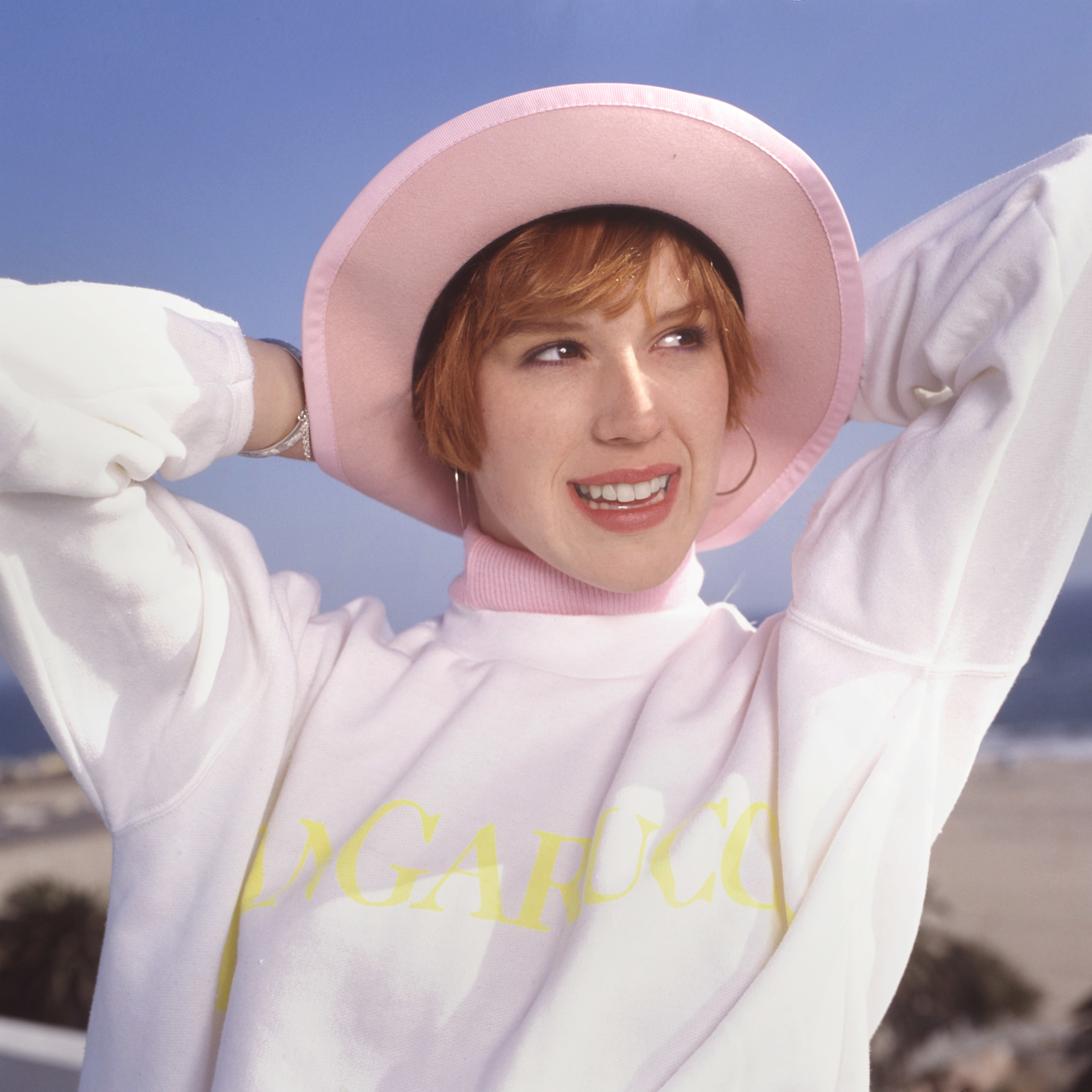 Molly Ringwald in a pink hat and white sweatshirt with text on it, smiling against a sky backdrop