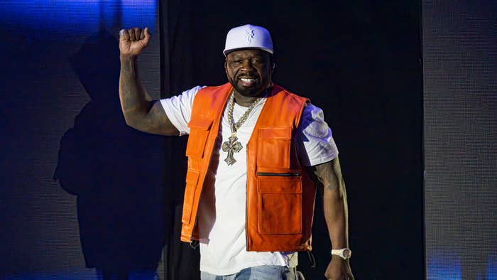 Person on stage in a white cap, orange vest over a white shirt, and gold chain