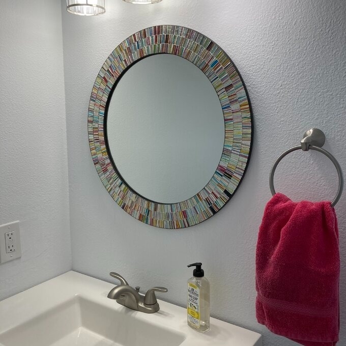 Round mirror with unique multicolored frame above a bathroom sink, next to a towel holder with a hanging towel