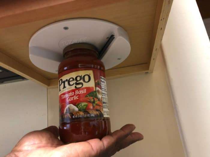 A person&#x27;s hand supporting a jar of Prego Tomato Basil Garlic sauce that&#x27;s stuck to an underside cabinet shelf, demonstrating a storage solution