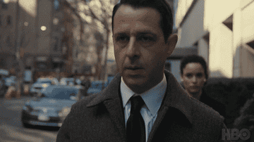 Two actors walk confidently, the man in a coat and the woman in a blazer, in an urban setting