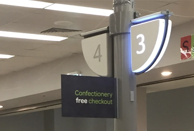 Checkout lanes in a store with a &quot;Confectionery free checkout&quot; sign, with shoppers and a baby visible