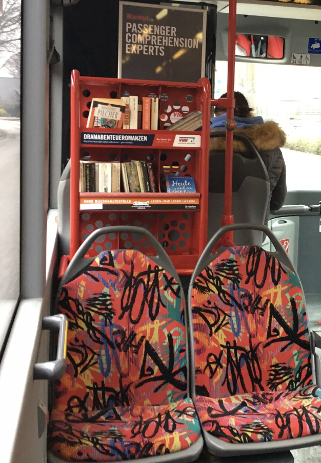 Bus interior with a bookshelf above two patterned seats, encouraging reading during travel
