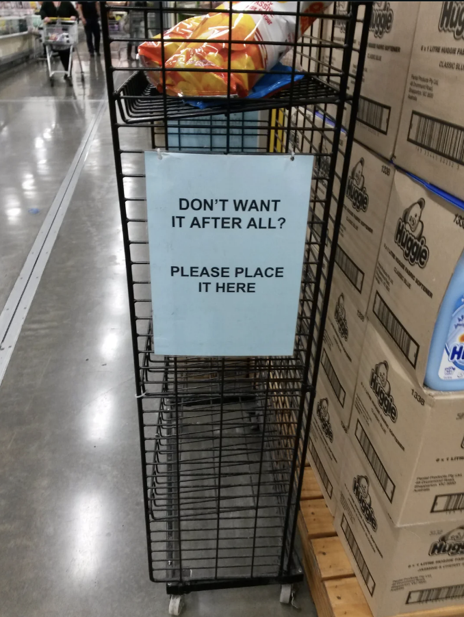 A sign in a store reads &quot;DON&#x27;T WANT IT AFTER ALL? PLEASE PLACE IT HERE&quot; on a metal rack with items returned by shoppers