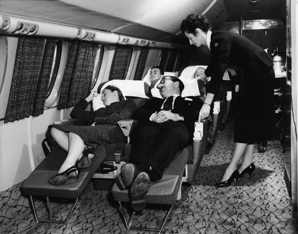 Black and white photo of passengers in vintage aircraft sleeper seats with an attendant standing by