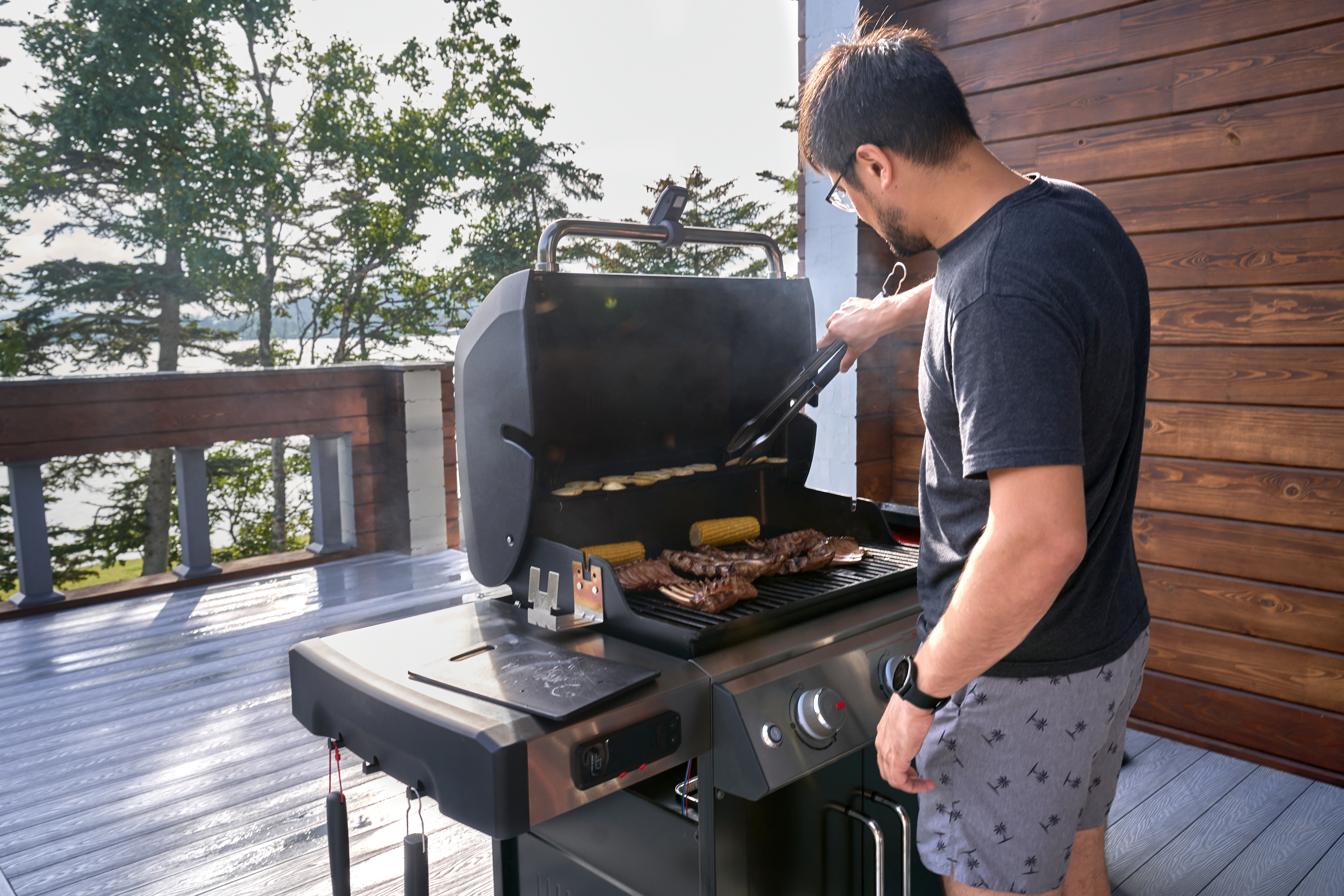 Person grilling food on an outdoor barbecue on a deck, with trees and a lake in the background