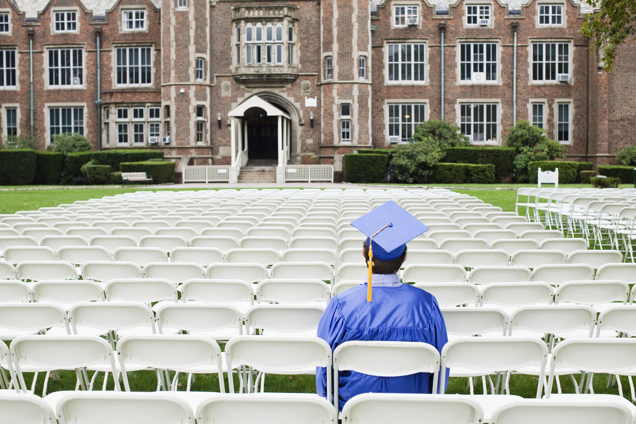 Graduate in blue cap and gown standing in front of rows of empty chairs facing a stage, building in background