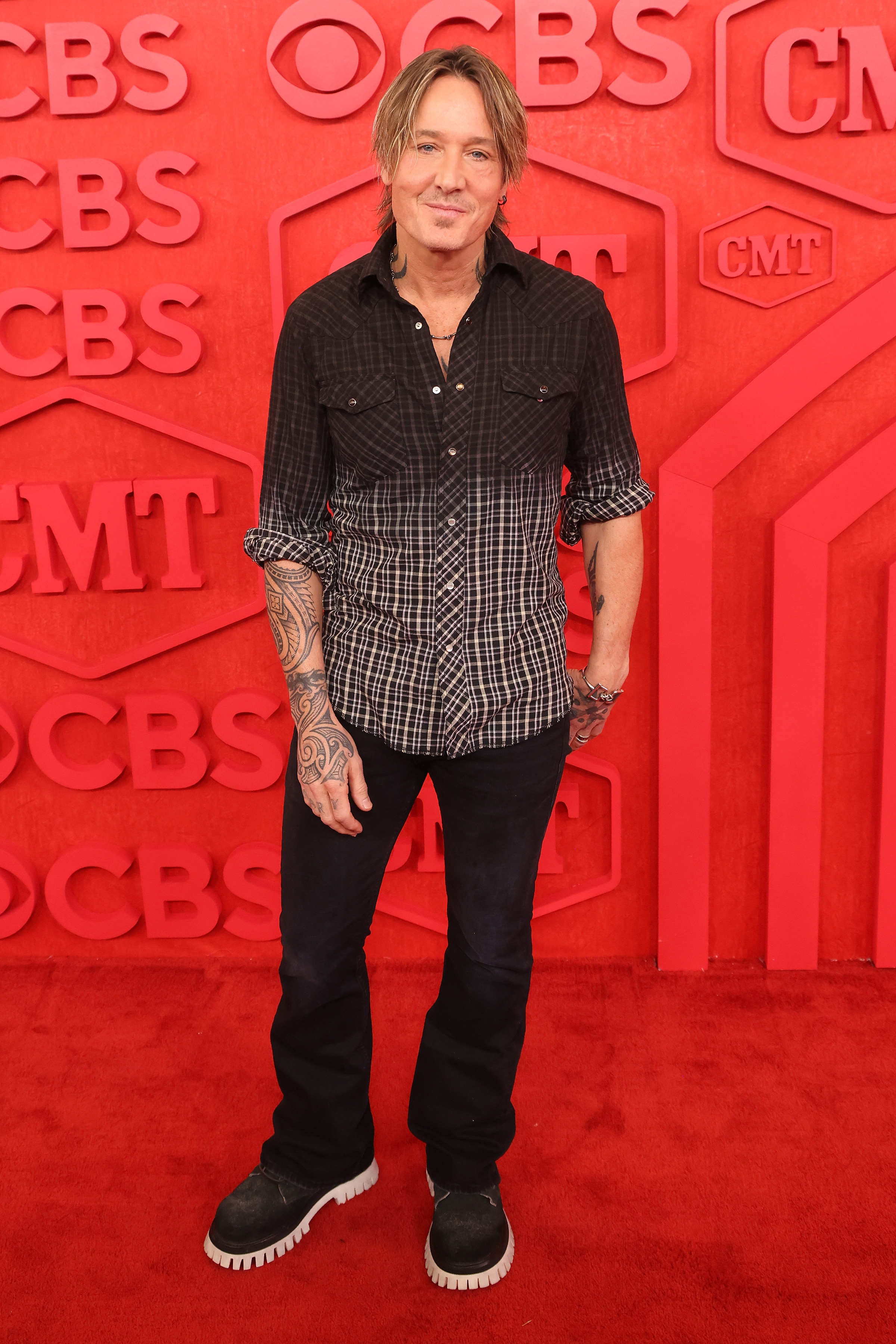 Keith Urban in a black and white checkered shirt with rolled-up sleeves and jeans, standing at a red carpet event