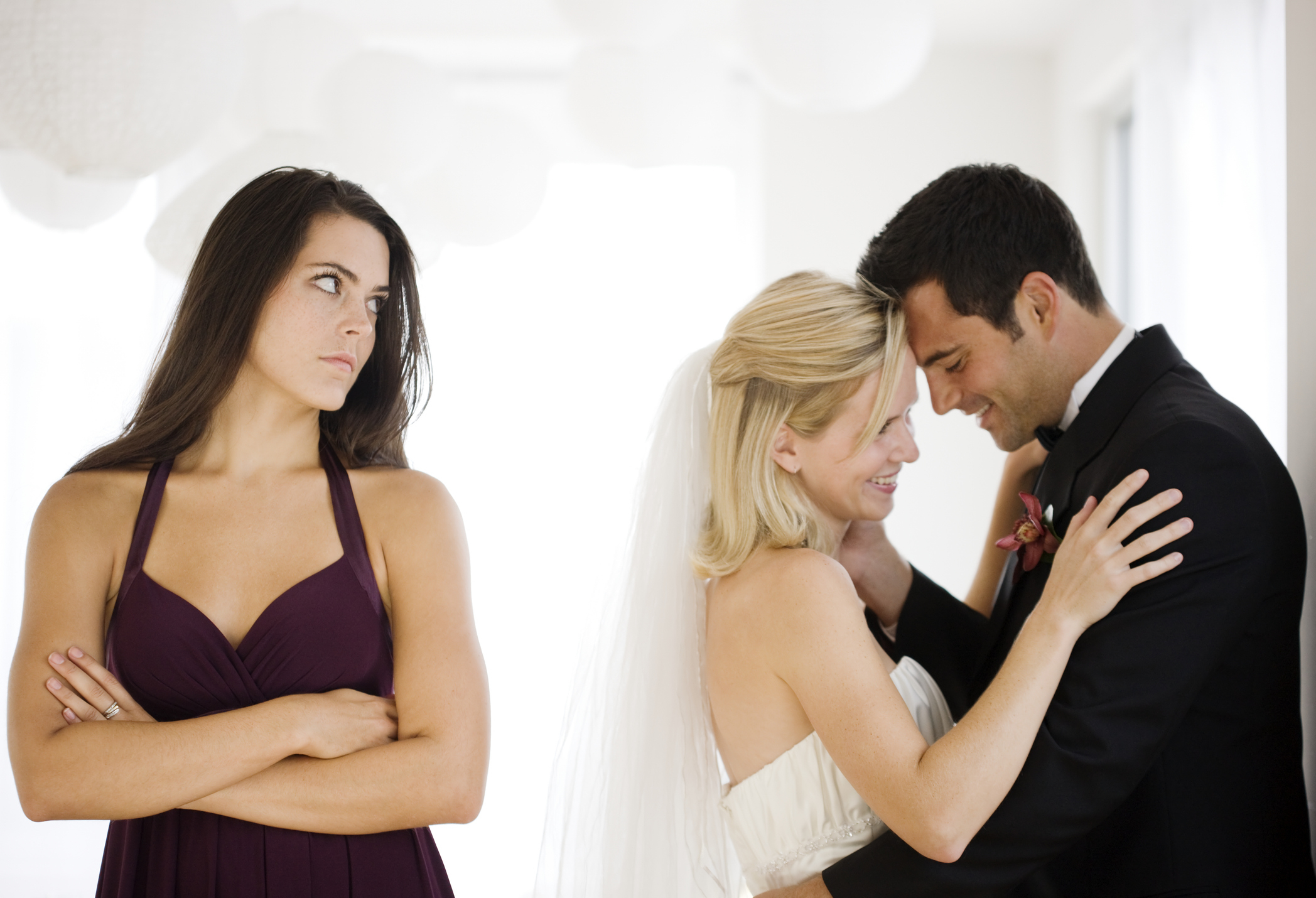 Woman in bridesmaid dress looks on angrily as bride and groom embrace