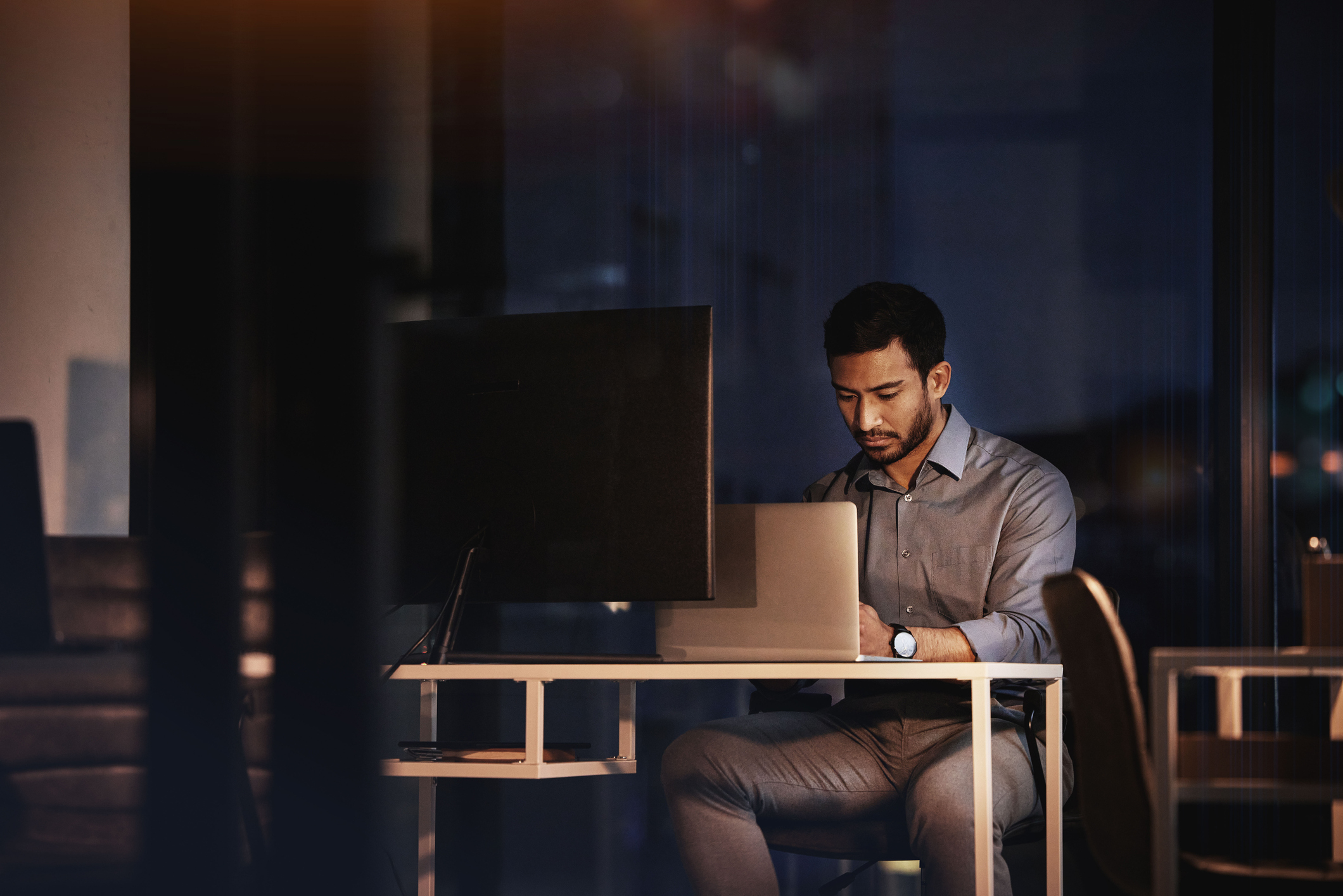 Man in a shirt working at a desk with a laptop and monitor at night