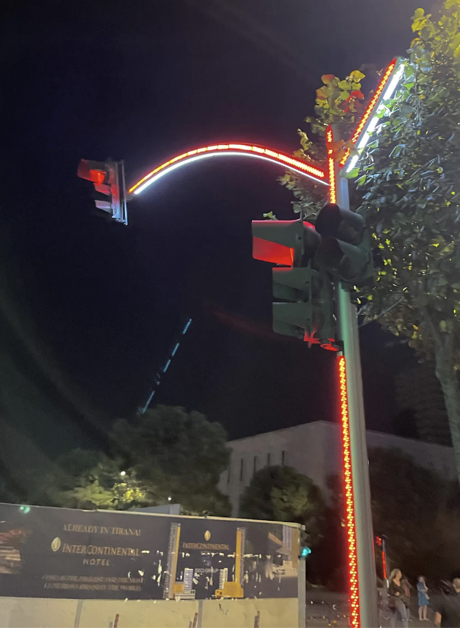 Street view at night with neon-lit arch, traffic light, and hotel signage. People are present near a bus stop