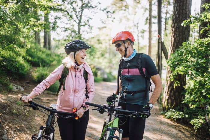 Two people, wearing helmets and casual outdoor attire, are standing with bicycles on a forest trail, smiling at each other