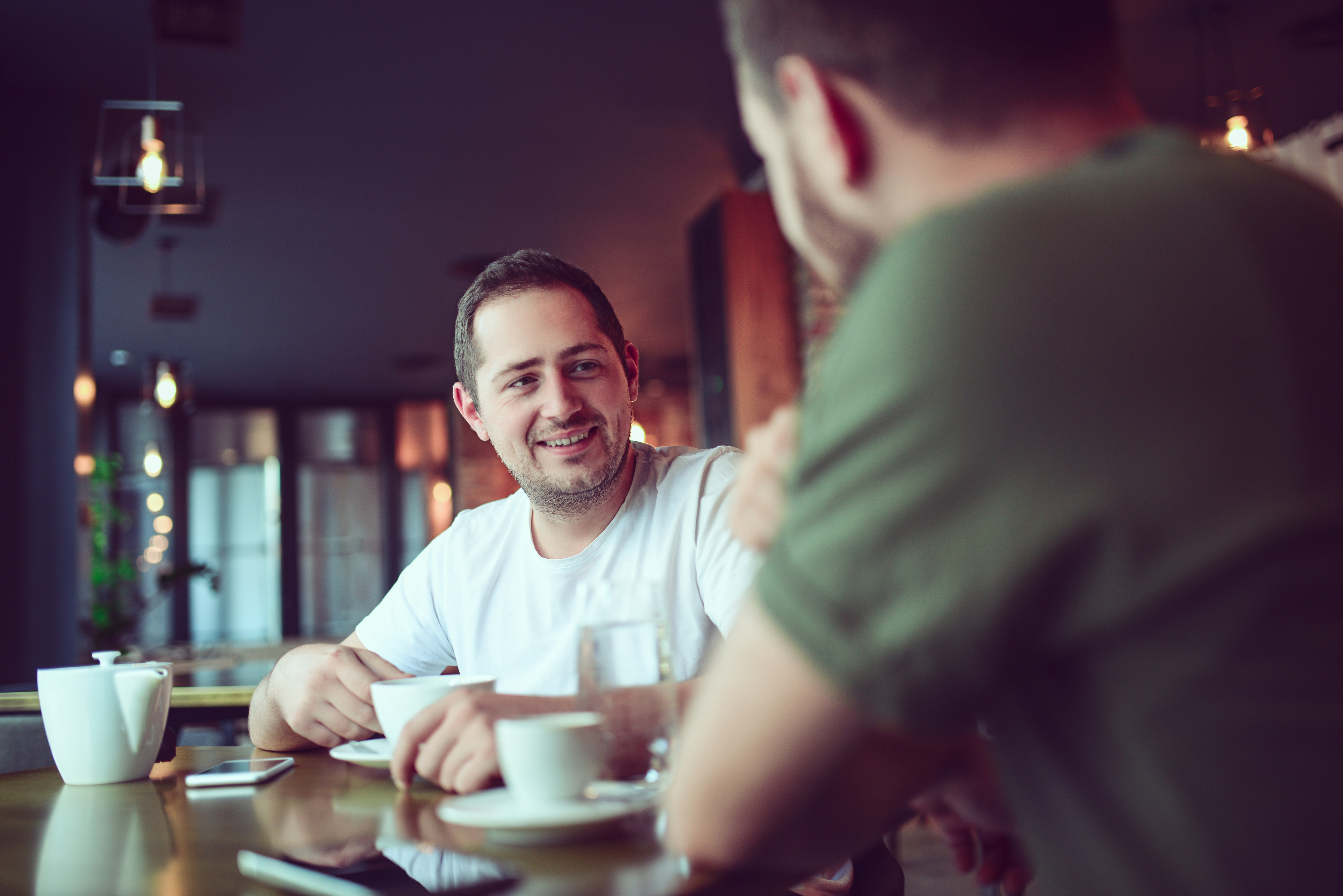 Two people having a conversation at a cafe table with cups in front of them. One is facing the camera smiling