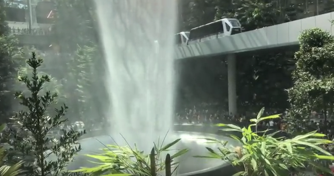 Waterfall cascading from a high bridge into a park with spectators and a bus on top of the bridge