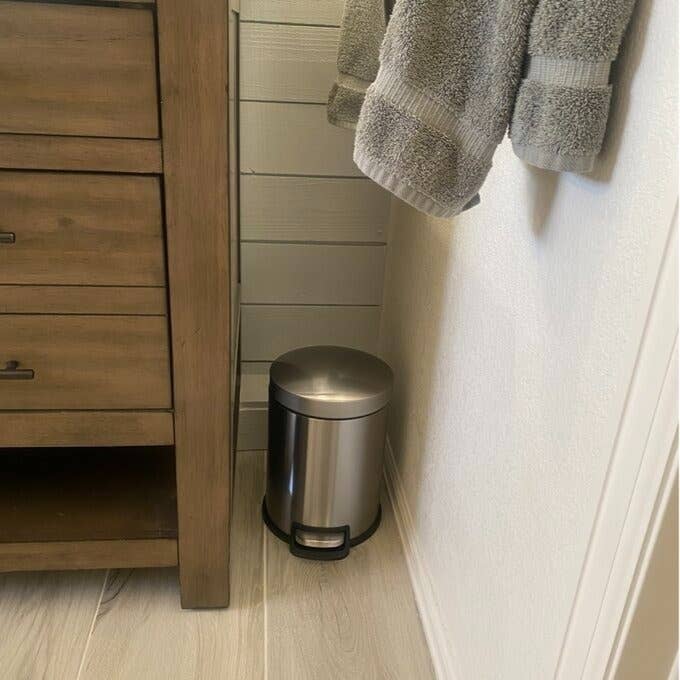 A stainless steel pedal trash can next to a wooden vanity