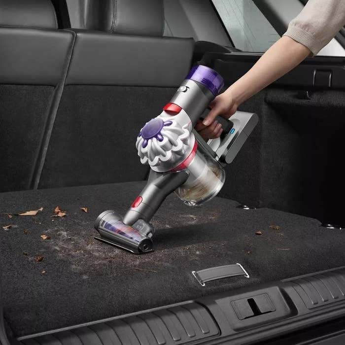 A handheld vacuum cleaner is being used to clean debris from a car&#x27;s rear seat floor