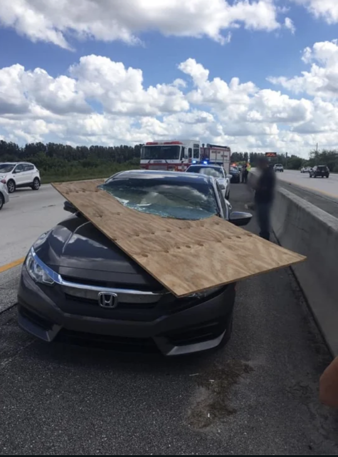 Car on highway with large wooden board crashed through its windshield; emergency vehicles in the background