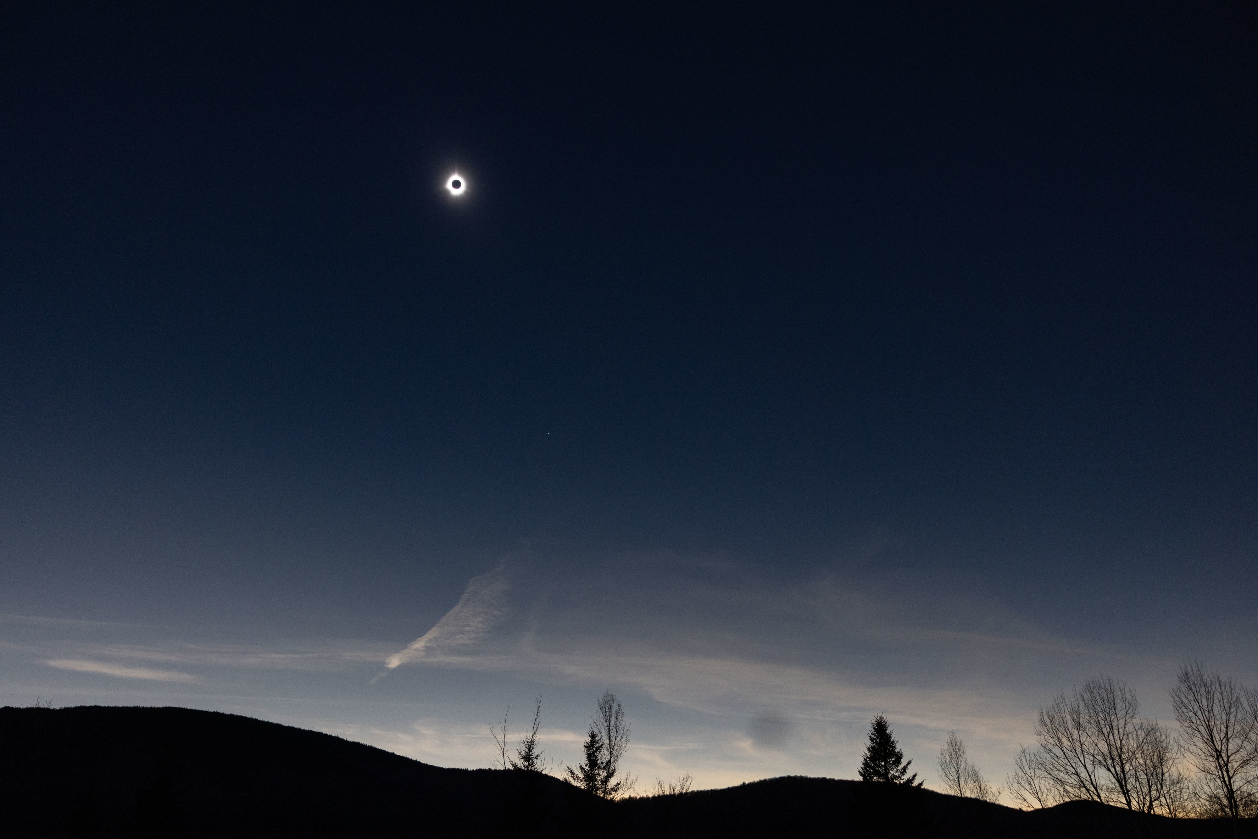 Total solar eclipse with corona visible over silhouette of hills and sparse foliage