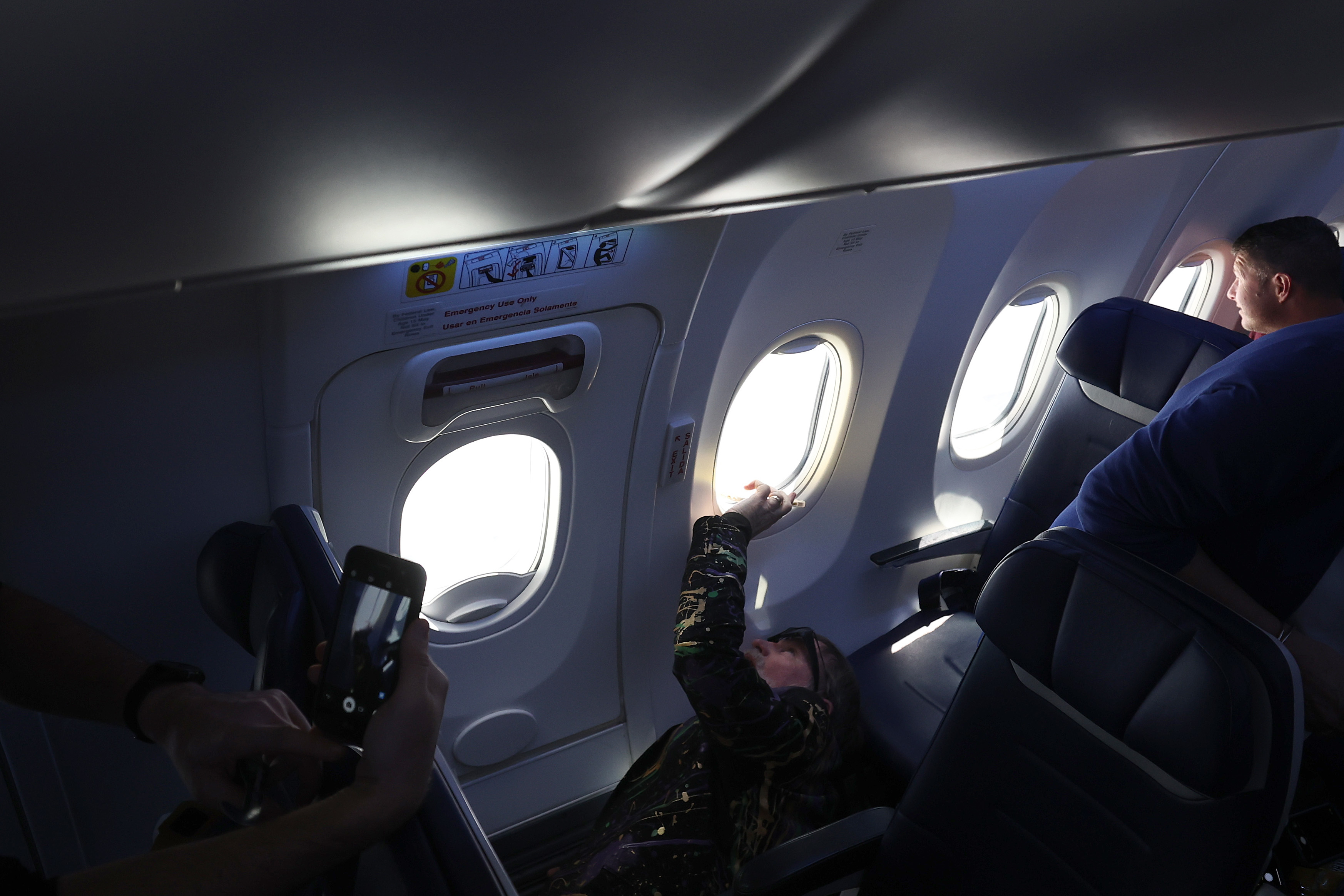 Passengers on a plane with one person pulling down a window shade; others record the event with phones