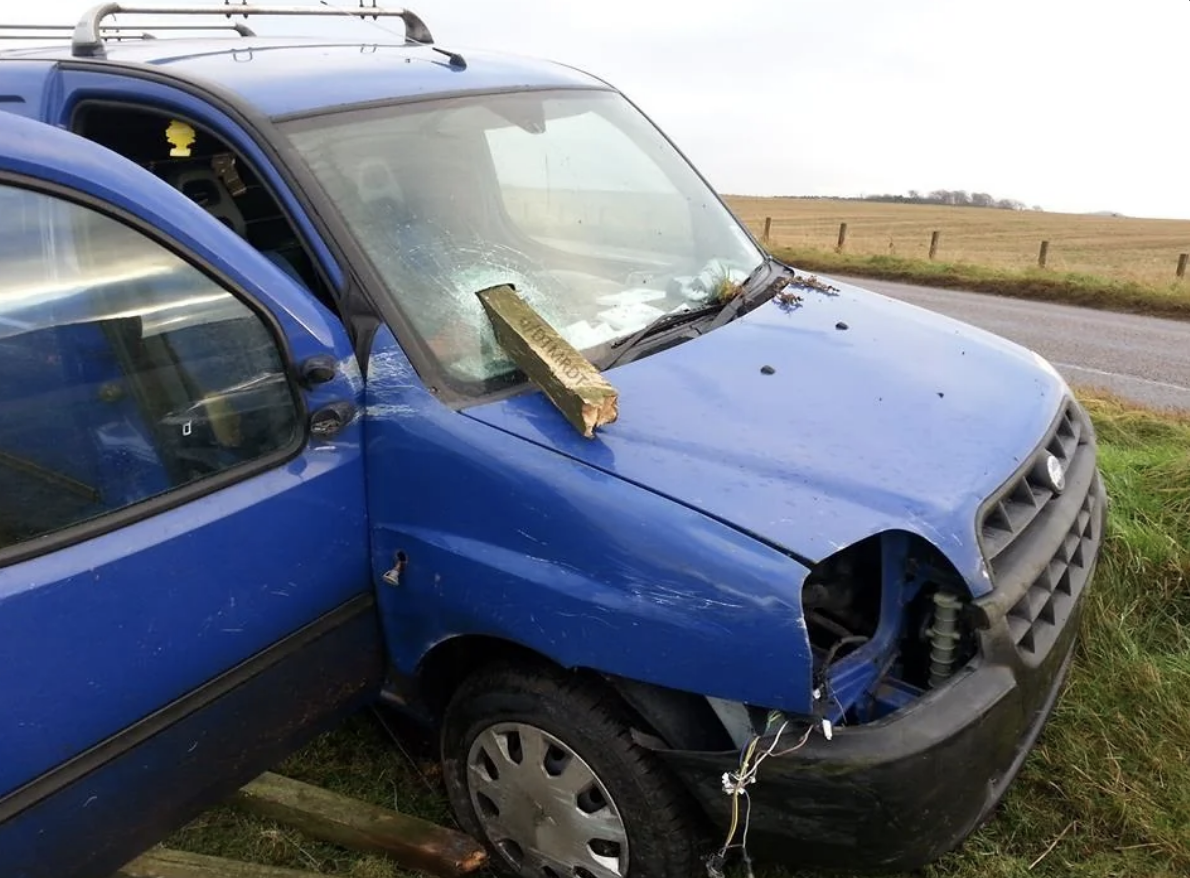 Damaged blue car with a wooden plank through the windshield, parked on grass beside a road