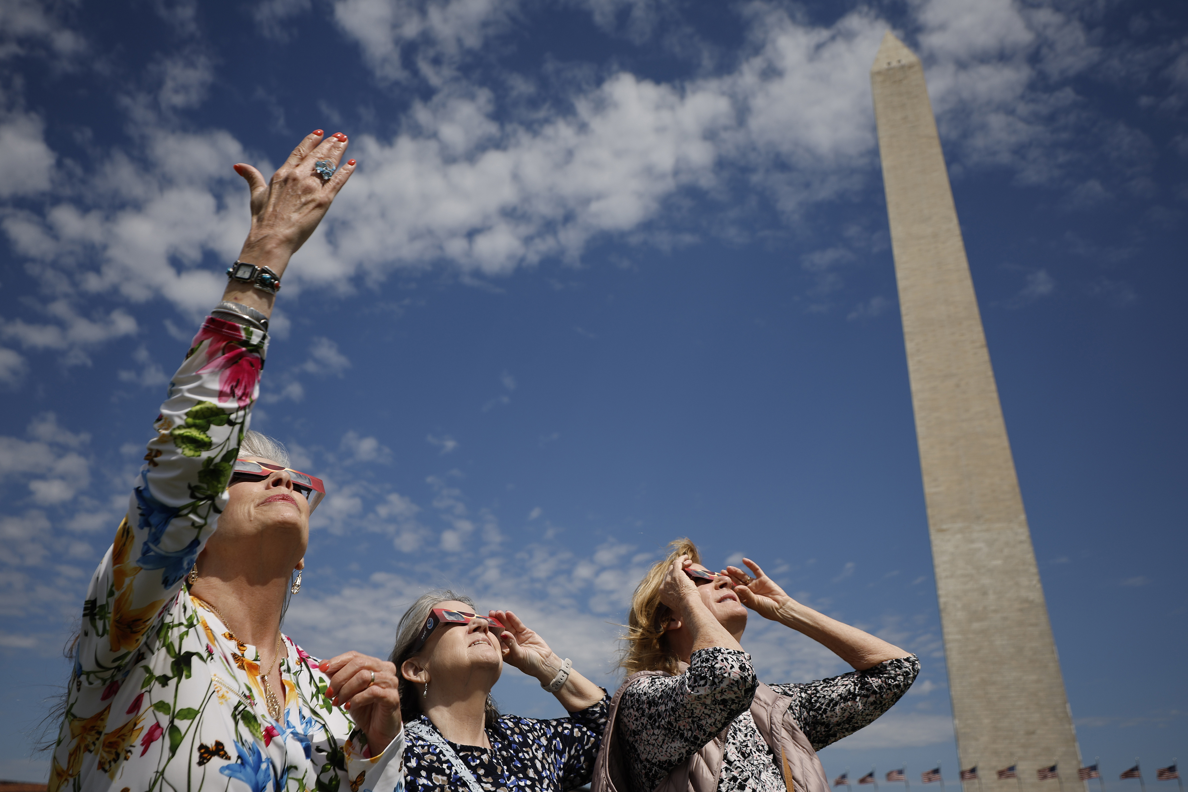 Three people looking upwards with hands raised near the Washington Monument, clear sky in the background