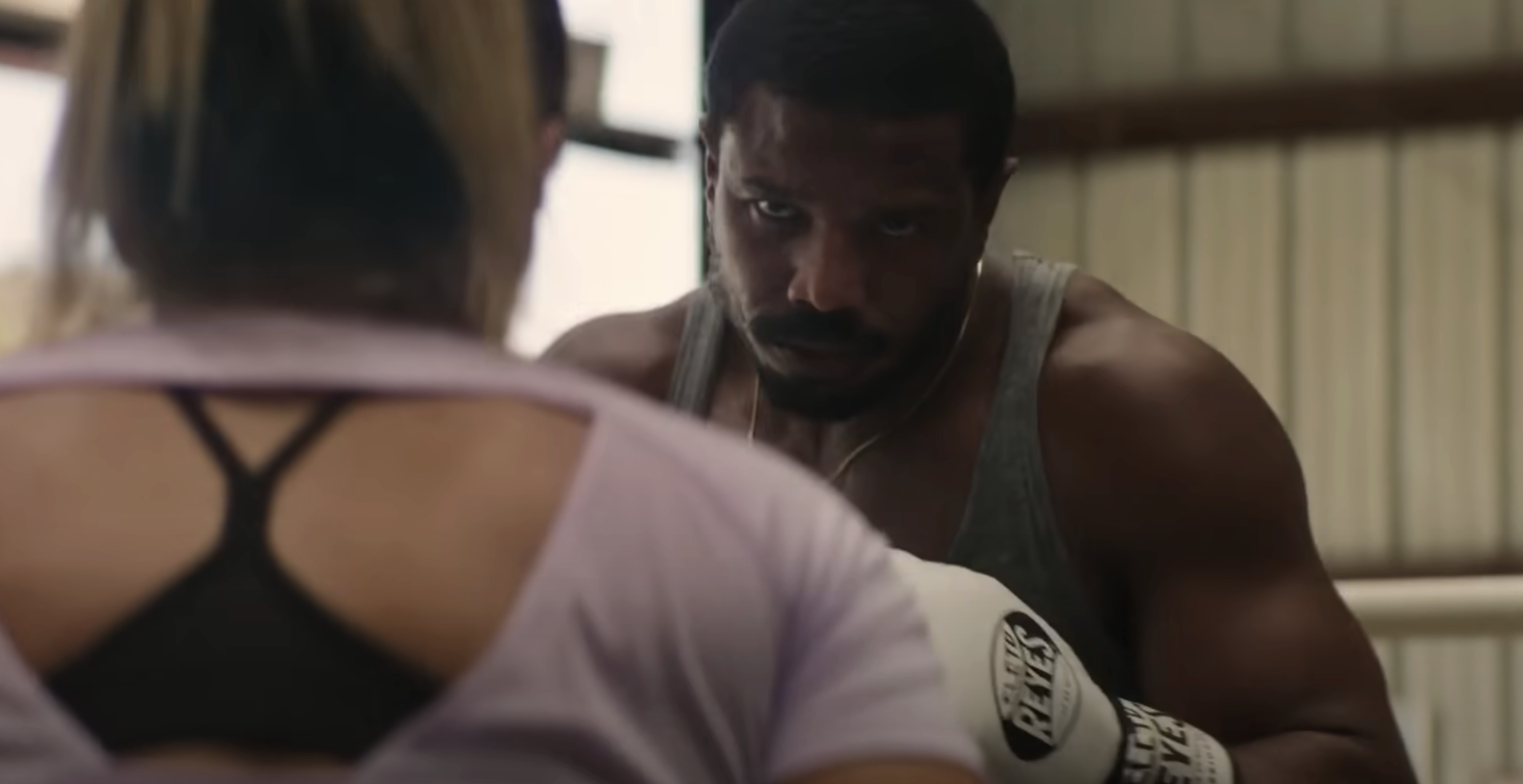Michael B. Jordan in a gym, wearing boxing gloves, facing a woman with her back to the camera