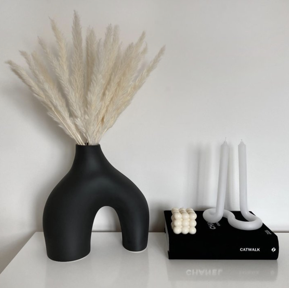 Sculptural vase with pampas grass next to a candle holder with white candles, both on a shelf