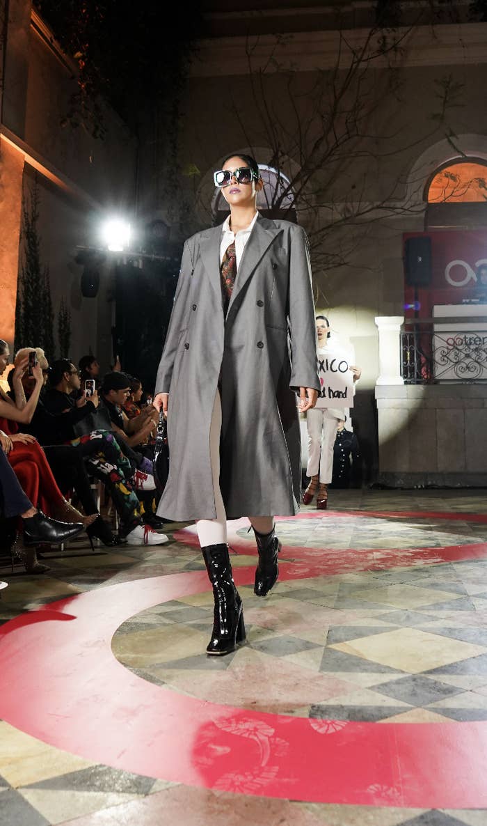Modelo en pasarela con gabardina gris, botas altas negras, gafas de sol y corbata, en evento de moda