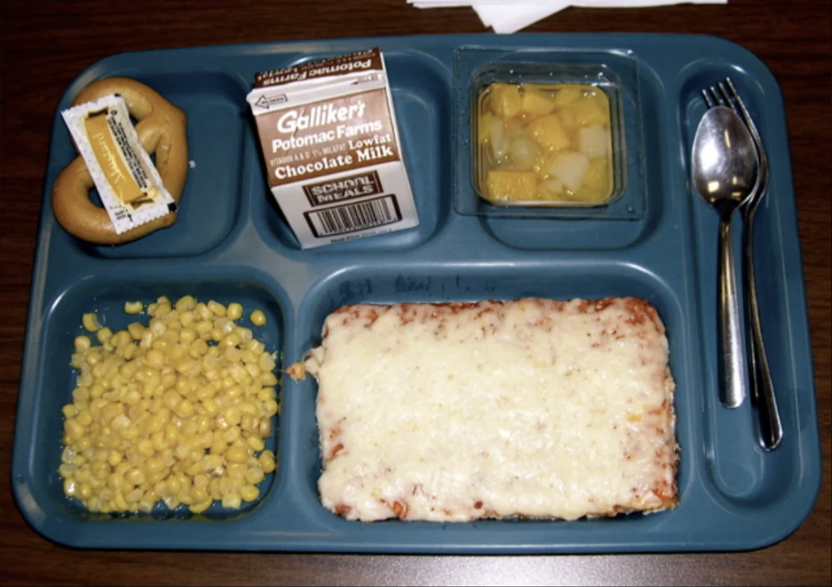 School lunch tray with corn, cheese pizza, pineapples, chocolate milk, and a bread roll